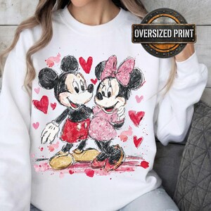 May include: White sweatshirt featuring a watercolor illustration of Mickey and Minnie Mouse embracing, surrounded by red hearts. Mickey wears red shorts, and Minnie wears a pink dress and bow. The sweatshirt has an "Oversized Print" label.