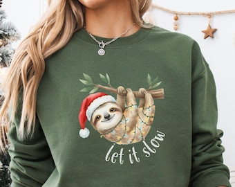 May include: Military green sweatshirt featuring a festive sloth wearing a Santa hat and string lights, hanging from a branch. The words "let it slow" are written in white script below the sloth. The sweatshirt is a comfortable, long-sleeved design.