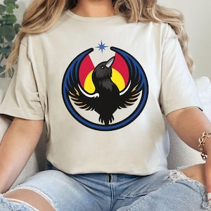 May include: Sand-colored t-shirt featuring a circular graphic of a black raven with outstretched wings, set against a backdrop of red, yellow, and blue. The raven is encircled by a blue ring, with a star above its head.