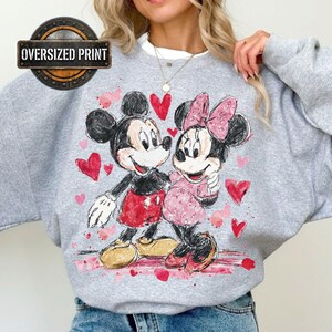 A person wearing a comfortable sweatshirt featuring an oversized retro-style watercolor print of Mickey and Minnie Mouse looking at each other, surrounded by scattered red and pink hearts.