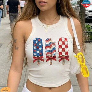 May include: A white vest top with three popsicle designs in red, white, and blue. The first popsicle has blue stars, the second has red and white stripes, and the third has a red and white checkerboard pattern. Each popsicle has a red bow.