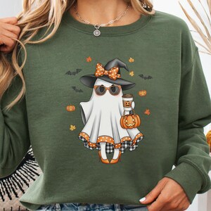 May include: A military green crewneck sweatshirt featuring a graphic of a white ghost wearing a black witch hat with an orange polka dot bow, sunglasses, and orange heels. The ghost holds a coffee cup and a pumpkin, surrounded by bats and leaves.