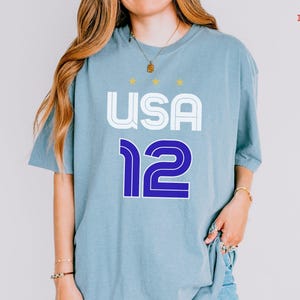 K&ouml;nnte beinhalten: Eisblaues T-Shirt mit dem Wort "USA" in Wei&szlig; und der Zahl "12" in Blau. Das Shirt hat einen Rundhalsausschnitt und kurze &Auml;rmel. &Uuml;ber dem Wort "USA" befinden sich drei kleine goldene Sterne.