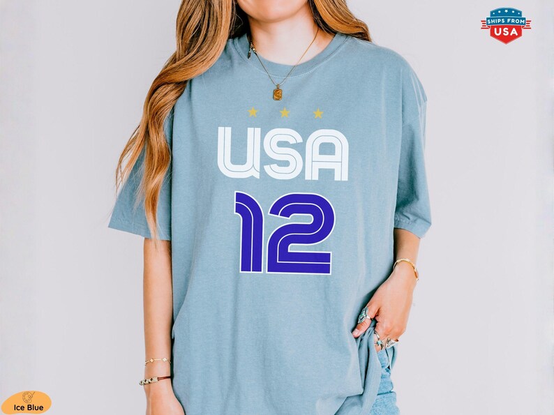 K&ouml;nnte beinhalten: Eisblaues T-Shirt mit dem Wort "USA" in Wei&szlig; und der Zahl "12" in Blau. Das Shirt hat einen Rundhalsausschnitt und kurze &Auml;rmel. &Uuml;ber dem Wort "USA" befinden sich drei kleine goldene Sterne.