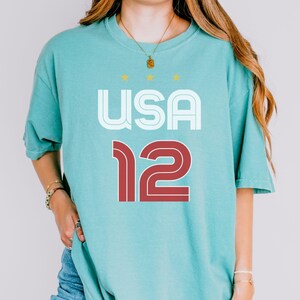 K&ouml;nnte beinhalten: Meergr&uuml;nes T-Shirt mit dem Wort "USA" in Wei&szlig; und der Zahl "12" in Rot. Das Shirt hat eine lockere Passform und kurze &Auml;rmel. &Uuml;ber dem Wort "USA" befinden sich drei gelbe Sterne.