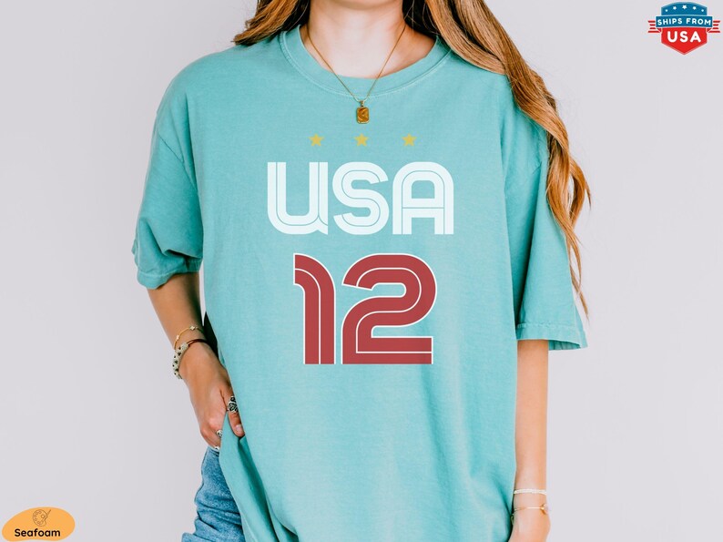 K&ouml;nnte beinhalten: Meergr&uuml;nes T-Shirt mit dem Wort "USA" in Wei&szlig; und der Zahl "12" in Rot. Das Shirt hat eine lockere Passform und kurze &Auml;rmel. &Uuml;ber dem Wort "USA" befinden sich drei gelbe Sterne.