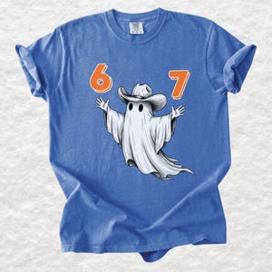 May include: A blue short-sleeved t-shirt featuring a graphic of a white ghost wearing a cowboy hat with its arms outstretched. The numbers 6 and 7 are printed in orange above the ghost. The shirt has a crew neckline and a casual, relaxed fit.