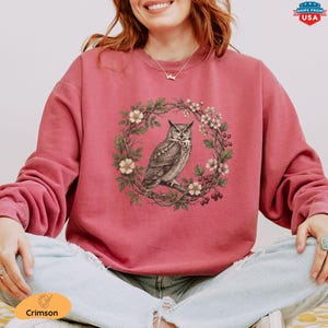 May include: Crimson-colored sweatshirt featuring a detailed illustration of an owl perched on a branch within a floral wreath. The owl is depicted in shades of brown and gray, surrounded by white flowers and green leaves. The sweatshirt has long sleeves.
