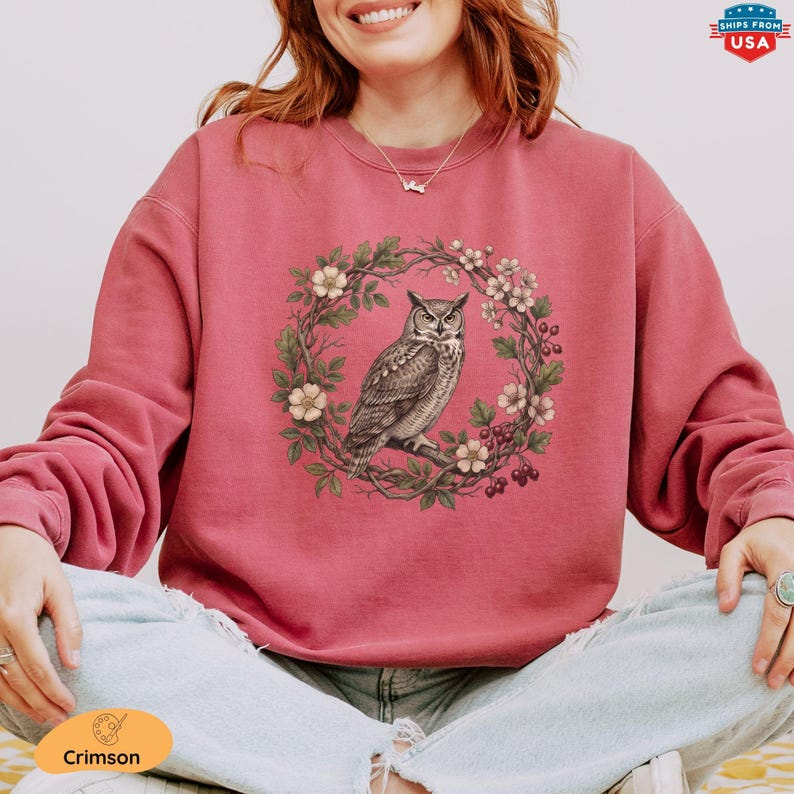 May include: Crimson-colored sweatshirt featuring a detailed illustration of an owl perched on a branch within a floral wreath. The owl is depicted in shades of brown and gray, surrounded by white flowers and green leaves. The sweatshirt has long sleeves.