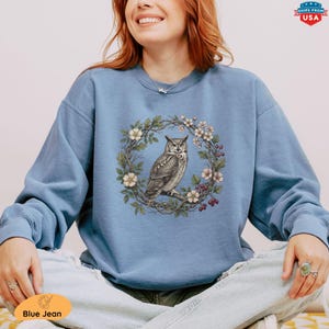 May include: A light blue sweatshirt featuring a detailed illustration of an owl perched on a branch within a floral wreath. The design includes white flowers, green leaves, and red berries. The text "Blue Jean" is on the bottom left.