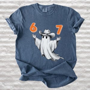 May include: A denim blue short-sleeve t-shirt featuring a graphic of a white ghost wearing a cowboy hat. The ghost is positioned between the orange numbers 6 and 7. The design is centered on the chest of the shirt, which has a crew neckline.