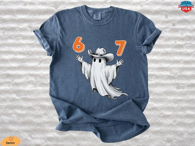 May include: A denim blue short-sleeve t-shirt featuring a graphic of a white ghost wearing a cowboy hat. The ghost is positioned between the orange numbers 6 and 7. The design is centered on the chest of the shirt, which has a crew neckline.