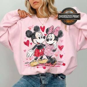 Op de afbeelding: Roze oversized print sweatshirt met een aquarel illustratie van Mickey en Minnie Mouse, omringd door rode harten. Het ontwerp bevat de tekst "Oversized Print" in een cirkelvormig kader.