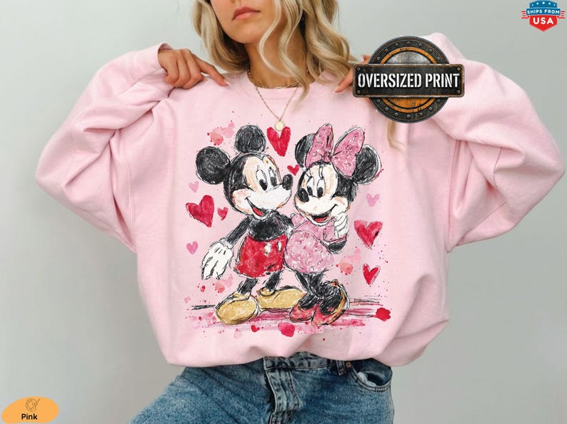 Op de afbeelding: Roze oversized print sweatshirt met een aquarel illustratie van Mickey en Minnie Mouse, omringd door rode harten. Het ontwerp bevat de tekst "Oversized Print" in een cirkelvormig kader.