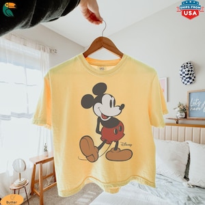 K&ouml;nnte beinhalten: Gelbes T-Shirt mit einer Grafik von Mickey Mouse in einer Laufpose. Die Disney-Figur ist mit schwarzen Ohren, einem wei&szlig;en Gesicht, roten Shorts und braunen Schuhen dargestellt. Das Shirt h&auml;ngt an einem Holzb&uuml;gel.