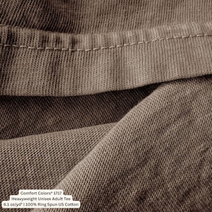 May include: Close-up of a brown heavyweight unisex adult tee shirt. The fabric is folded, showing the texture and stitching details. The text on the image reads: Comfort Colors 1717, Heavyweight Unisex Adult Tee, 6.1 oz/yd&sup2; | 100% Ring Spun US Cotton.