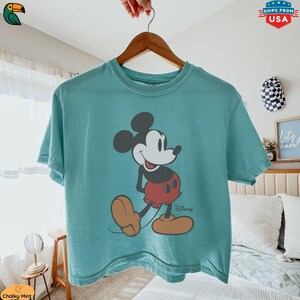 K&ouml;nnte beinhalten: Ein t&uuml;rkisfarbenes T-Shirt mit einer klassischen Mickey Mouse-Grafik. Die Zeichentrickfigur ist in Schwarz, Wei&szlig;, Rot und Beige dargestellt, mit dem Disney-Logo darunter. Das T-Shirt h&auml;ngt an einem Holzb&uuml;gel.