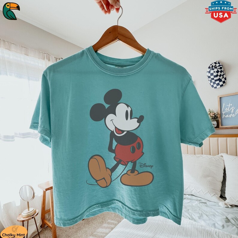 K&ouml;nnte beinhalten: Ein t&uuml;rkisfarbenes T-Shirt mit einer klassischen Mickey Mouse-Grafik. Die Zeichentrickfigur ist in Schwarz, Wei&szlig;, Rot und Beige dargestellt, mit dem Disney-Logo darunter. Das T-Shirt h&auml;ngt an einem Holzb&uuml;gel.