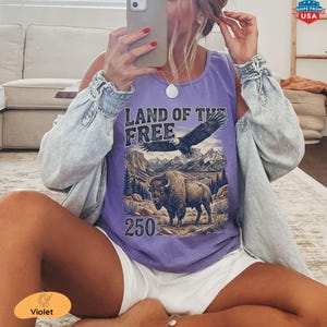 May include: A violet tank top with the text "LAND OF THE FREE" and an illustration of an eagle, bison, and mountains. The number "250" is below the illustration. The tank top is paired with white shorts and a light blue denim jacket.