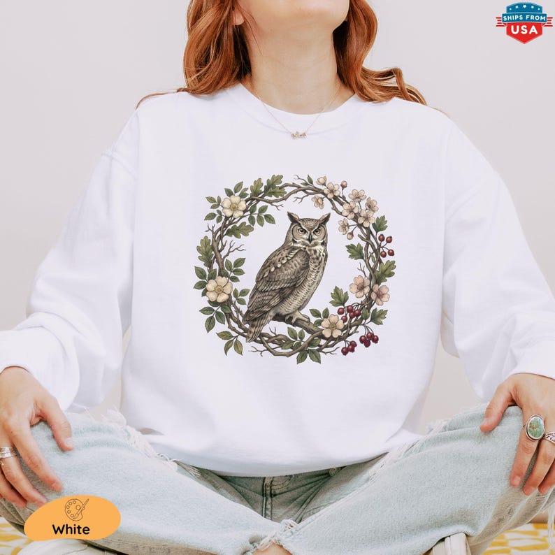 May include: White sweatshirt featuring a detailed illustration of an owl perched on a branch within a floral wreath. The owl is depicted in shades of brown and gray, surrounded by green leaves, white and pink flowers, and red berries. The design is centered on the front.
