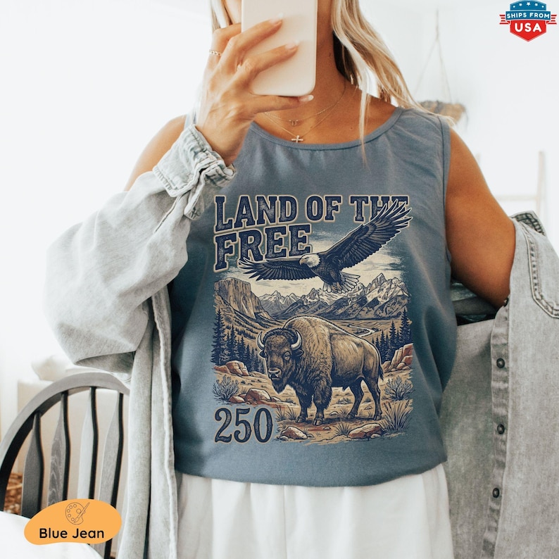 May include: Blue tank top with a graphic featuring an eagle, bison, mountains, and the text "LAND OF THE FREE 250". The design is in shades of brown, blue, and white. The tank top is a blue jean color.