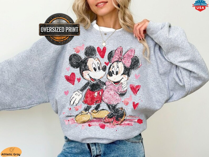 A person wearing a comfortable sweatshirt featuring an oversized retro-style watercolor print of Mickey and Minnie Mouse looking at each other, surrounded by scattered red and pink hearts.
