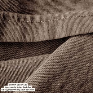 May include: A brown t-shirt featuring a visible hemline and a textured fabric weave. The garment is labeled as a Comfort Colors 1717 Heavyweight Unisex Adult Tee, made of 6.1 oz/yd&sup2; 100% Ring Spun US Cotton, as indicated by the printed text.
