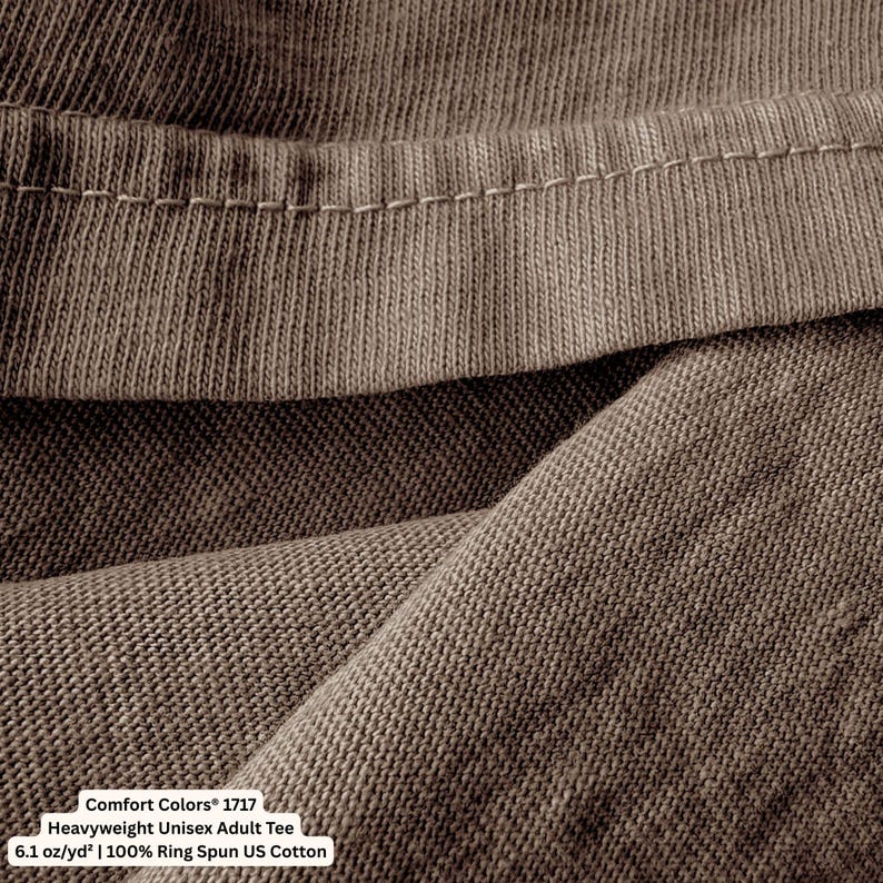 May include: A brown t-shirt featuring a visible hemline and a textured fabric weave. The garment is labeled as a Comfort Colors 1717 Heavyweight Unisex Adult Tee, made of 6.1 oz/yd&sup2; 100% Ring Spun US Cotton, as indicated by the printed text.