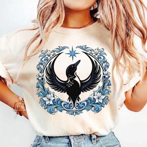 May include: Ivory-colored t-shirt featuring a black bird with outstretched wings, a red eye, and a star above. The bird is encircled by a blue and white floral design. The shirt is made of a soft material.