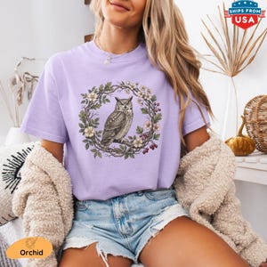 May include: A light purple t-shirt featuring a detailed illustration of an owl perched within a floral wreath. The design includes white flowers, green leaves, and red berries. The shirt is a soft, comfortable material.