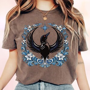 May include: Espresso-colored t-shirt featuring a black bird design with outstretched wings, a red eye, and a star above. The bird is encircled by a blue and white floral wreath. The shirt has a crew neck and short sleeves.
