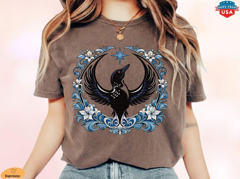 May include: Espresso-colored t-shirt featuring a black bird design with outstretched wings, a red eye, and a star above. The bird is encircled by a blue and white floral wreath. The shirt has a crew neck and short sleeves.