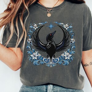 May include: Dark gray t-shirt featuring a black bird with outstretched wings, a star, and a floral wreath in blue and white. The design is centered on the shirt.