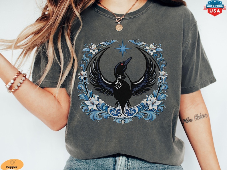 May include: Dark gray t-shirt featuring a black bird with outstretched wings, a star, and a floral wreath in blue and white. The design is centered on the shirt.