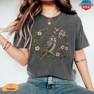 May include: Dark grey t-shirt featuring a detailed illustration of an owl perched within a floral wreath. The owl is depicted in shades of grey and brown, surrounded by white and pink flowers and red berries. The shirt has a vintage, worn look.