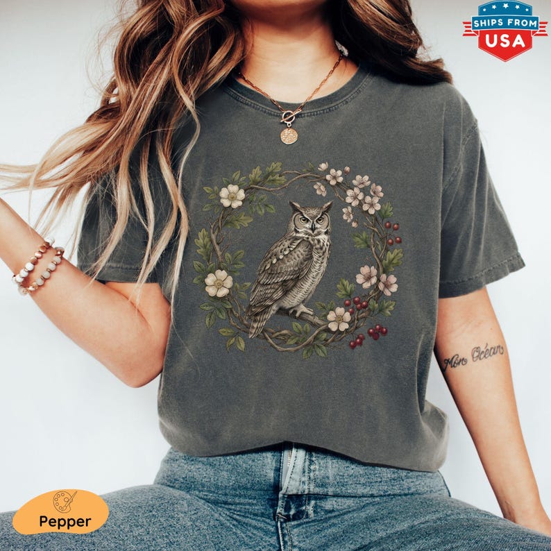 May include: Dark grey t-shirt featuring a detailed illustration of an owl perched within a floral wreath. The owl is depicted in shades of grey and brown, surrounded by white and pink flowers and red berries. The shirt has a vintage, worn look.