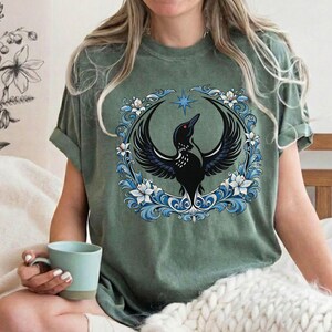 May include: Sage green t-shirt featuring a black bird with outstretched wings, set within a circular frame of blue and white floral elements. A blue star is above the bird. The shirt is a relaxed fit.