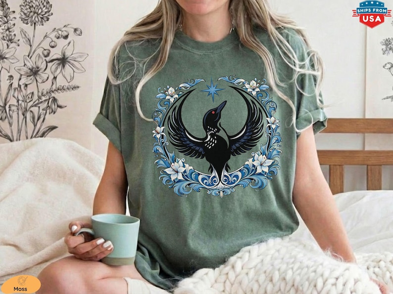 May include: Sage green t-shirt featuring a black bird with outstretched wings, set within a circular frame of blue and white floral elements. A blue star is above the bird. The shirt is a relaxed fit.