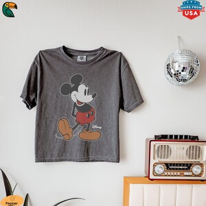 K&ouml;nnte beinhalten: Dunkelgraues T-Shirt mit einem Mickey Mouse-Motiv. Die Zeichentrickfigur ist in Schwarz, Rot und Beige dargestellt. Das T-Shirt hat einen Rundhalsausschnitt und kurze &Auml;rmel. Das Disney-Logo ist auf dem T-Shirt sichtbar.