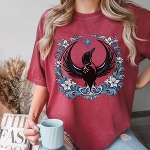 May include: Burgundy-colored t-shirt featuring a detailed graphic of a black bird with outstretched wings, encircled by a floral wreath with blue and white accents. A star is above the bird. The shirt is a comfortable, casual style.