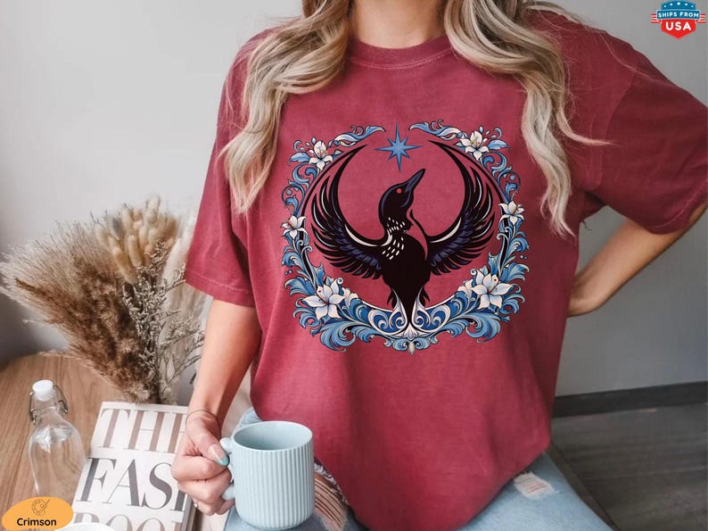 May include: Burgundy-colored t-shirt featuring a detailed graphic of a black bird with outstretched wings, encircled by a floral wreath with blue and white accents. A star is above the bird. The shirt is a comfortable, casual style.