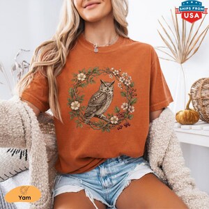 May include: Burnt orange t-shirt featuring a detailed illustration of an owl perched on a branch within a floral wreath. The design includes white flowers and red berries. The shirt is a comfortable, casual style.