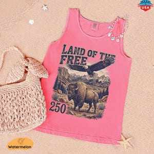 May include: A pink tank top with the text "LAND OF THE FREE" and a graphic of an eagle and bison. A woven handbag and a beaded necklace are next to the tank top. The tank top is on a sandy surface.