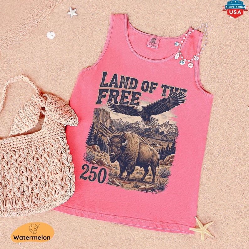 May include: A pink tank top with the text "LAND OF THE FREE" and a graphic of an eagle and bison. A woven handbag and a beaded necklace are next to the tank top. The tank top is on a sandy surface.