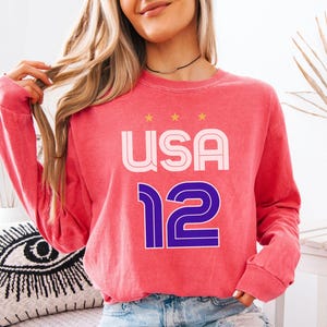 K&ouml;nnte beinhalten: Ein korallfarbenes Langarm-Sweatshirt mit dem Text "USA 12" in Wei&szlig; und Lila. Das Wort "USA" steht &uuml;ber der Zahl "12", mit drei goldenen Sternen &uuml;ber dem Wort "USA".