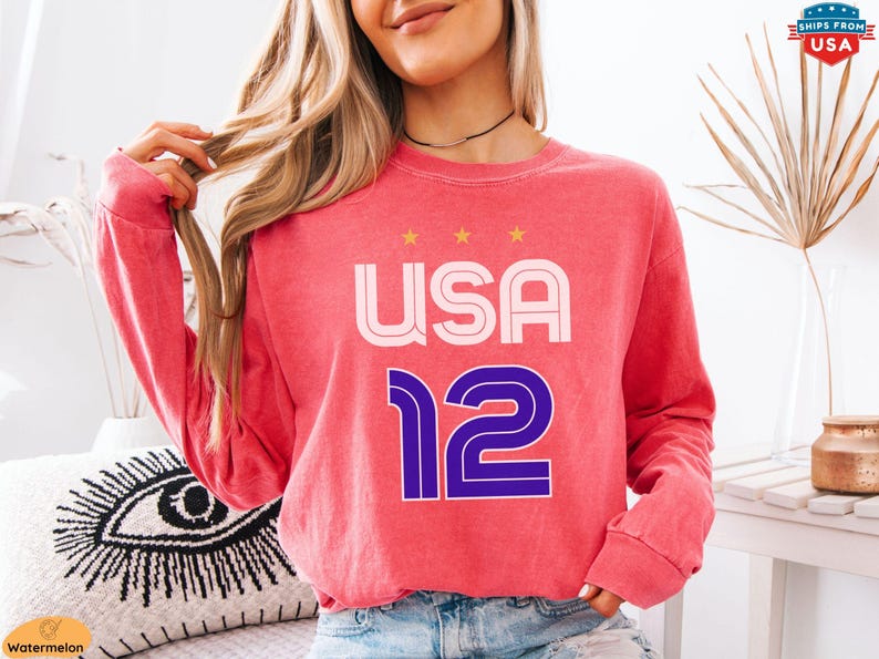 K&ouml;nnte beinhalten: Ein korallfarbenes Langarm-Sweatshirt mit dem Text "USA 12" in Wei&szlig; und Lila. Das Wort "USA" steht &uuml;ber der Zahl "12", mit drei goldenen Sternen &uuml;ber dem Wort "USA".