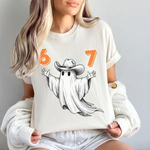 May include: An ivory short-sleeve t-shirt featuring a graphic of a white ghost wearing a brown cowboy hat with its arms outstretched. The number 6 is printed in orange to the left of the ghost, and the number 7 is printed in orange to the right.