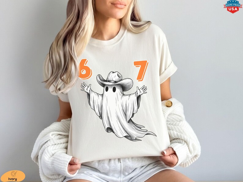 May include: An ivory short-sleeve t-shirt featuring a graphic of a white ghost wearing a brown cowboy hat with its arms outstretched. The number 6 is printed in orange to the left of the ghost, and the number 7 is printed in orange to the right.