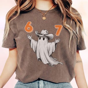 May include: An espresso-colored short-sleeve t-shirt featuring a graphic of a white ghost wearing a brown cowboy hat. The ghost is positioned between the orange numbers 6 and 7. The design is centered on the chest of the garment.