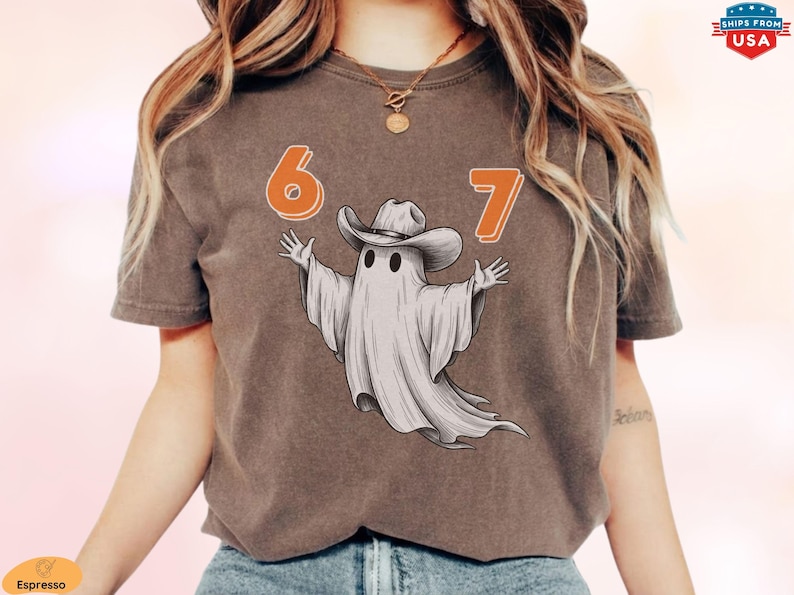 May include: An espresso-colored short-sleeve t-shirt featuring a graphic of a white ghost wearing a brown cowboy hat. The ghost is positioned between the orange numbers 6 and 7. The design is centered on the chest of the garment.